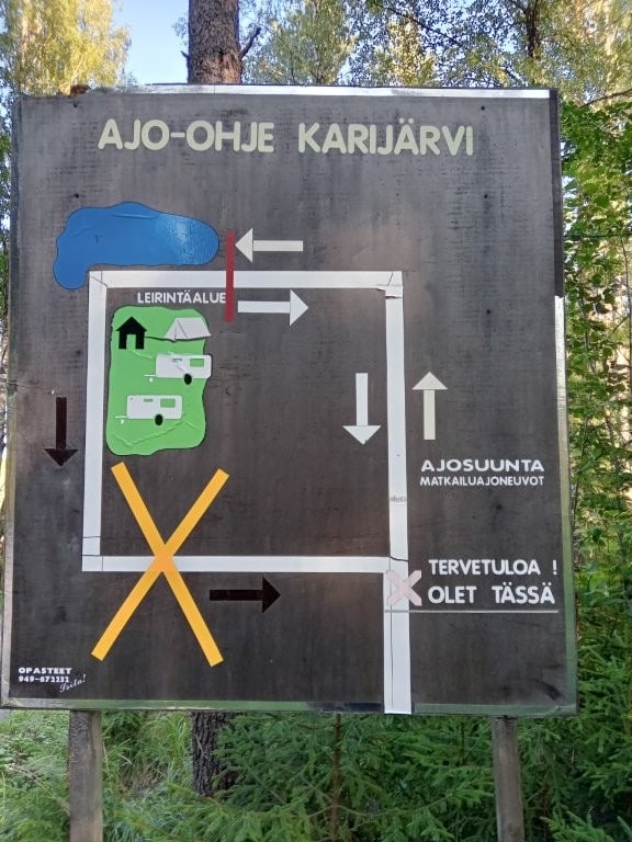 Käsin maalattu ajo-ohjetaulu: Ajo-ohje Karijärvi, kartta leirintäalueesta, keltainen rasti, mustat nuolet ja teksti Tervetuloa! Olet tässä.