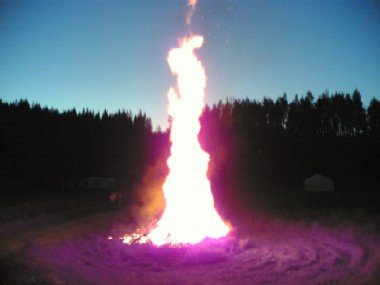 Korkea kokkotuli palaa hämärässä, taustalla asuntovaunuja ja metsän siluetti.