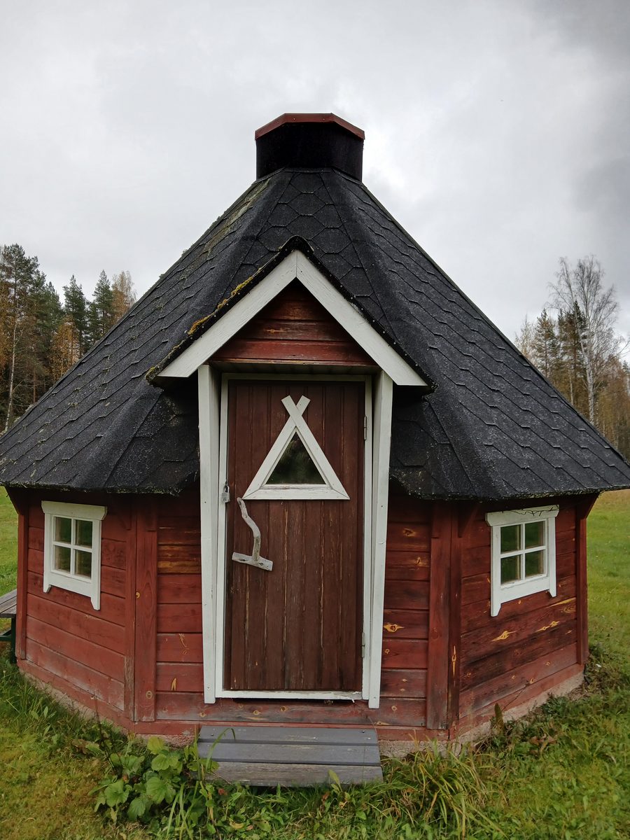 Punainen kahdeksankulmainen grillikota, musta paanukatto ja savupiippu, valkoiset ikkunanpielet ja ovessa kolmionmuotoinen ikkuna. Ympärillä nurmikkoa ja syksyistä metsää.